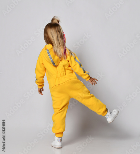 Blonde kid baby girlwith stylish hairstyle in modern yellow jumpsuit and sneakers stands back to camera on one leg