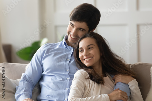 Harmony in relations. Happy affectionate millennial spouses or couple in love sitting on couch close together, embracing with warmth and looking aside watching nice movie on tv, creating future plans