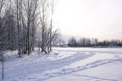 Winter landscape - white snow with traces of shoes and skis on the field. The ski track and road skirting the forest with bare trees, soft sunlight.