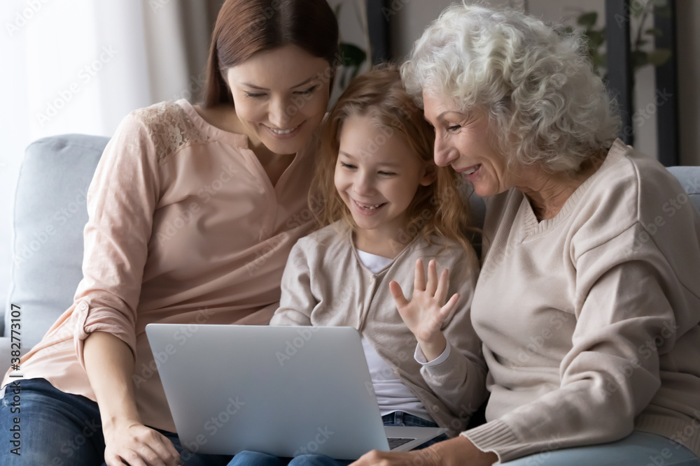 Happy little girl with mother and grandmother using laptop together, sitting on couch, looking at screen, family making video call, smiling pretty child waving hand, saying hello, greeting relatives Happy little girl with mother and grandmother using laptop together, sitting on couch, looking at screen, family making video call, smiling pretty child waving hand, saying hello, greeting relatives