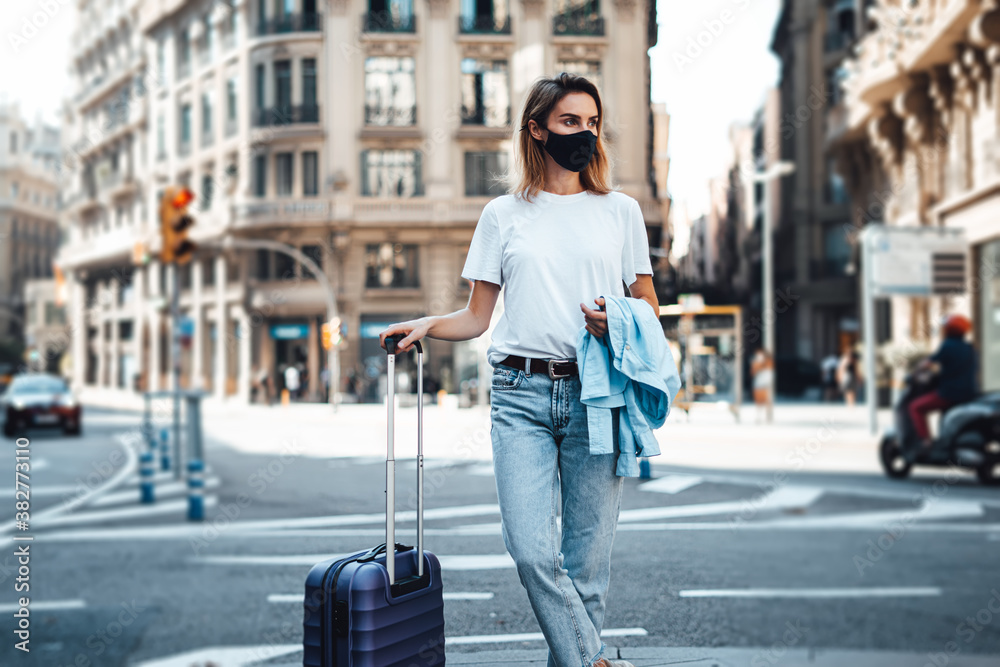 Young traveling woman with suitcase on a sunny city street. Traveler wearing mask on vacation. Young traveling woman with suitcase on a sunny city street. Traveler wearing mask on vacation.