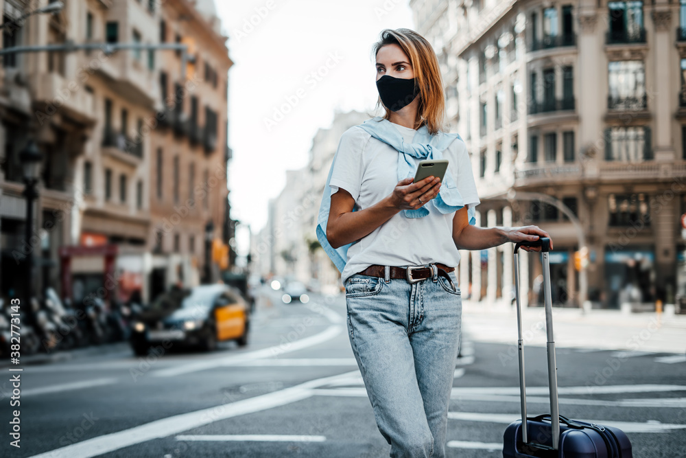 Obraz premium Young traveling woman with suitcase on a sunny city street. Traveler wearing mask on vacation.