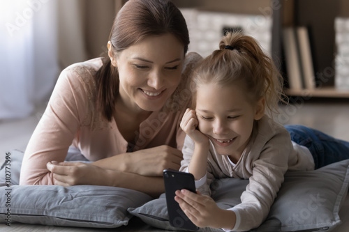 Happy little girl and young mother using phone together close up, lying on warm floor with underfloor heating, smiling woman with adorable girl enjoying leisure time, having fun with smartphone