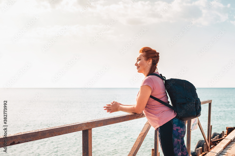 Portrait of a pretty adult woman In sportswear with a backpack posing smiling on the pier. In the background, the sea and sky. Side view. The concept of active recreation and freedom