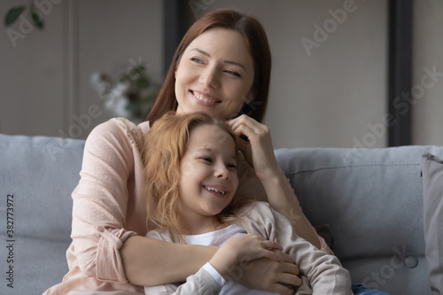 Smiling mother and adorable little daughter relaxing on couch together, beautiful young woman and pretty preschool girl child hugging, enjoying leisure time at home, having fun, cuddling