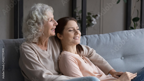 Smiling mature woman with grownup daughter relaxing on couch in living room, hugging, happy dreamy elderly female with granddaughter dreaming about good future, visualizing, enjoying leisure time