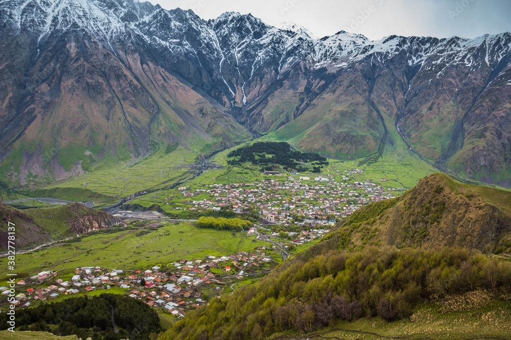 Naklejka premium Panoramic view on a small town Stepansminda, Georgia, in the downhills surrounded by Kazbek mountains in summer. Many small houses with rooftops in the valley, rural lifestyle in a village.