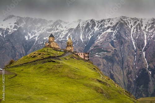 GERGETI, GEORGIA - 31 MAY, 2017. Panoramic view on Gergeti Trinity Church on the top of the hill against snowy mountains and white sky in summer.