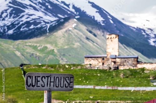 A wooden sign with the written word in English "Guesthouse" in the foreground. Medieval building on a green valley and snow capped mountains in the blurred background in Georgia. © Алексей Мараховец