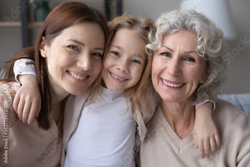 Head shot portrait smiling pretty little girl hugging young mother and mature grandmother, three generations of women looking at camera, posing for family photo, sitting on couch at home