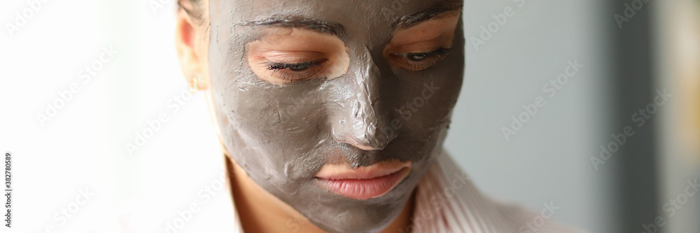 Fototapeta premium Close-up of young pretty female with clay mask on face. Attractive woman in pyjamas after night sleep. Copy space in left side. Morning facial treatment and relax concept