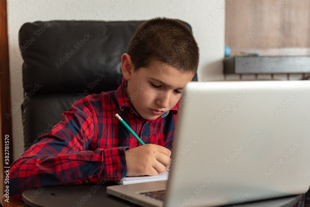 Young boy studying; watching online school program on laptop; home schooling; taking notes Young boy studying; watching online school program on laptop; home schooling; taking notes