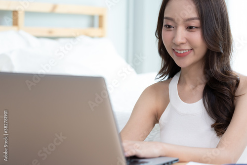 Asian woman working on a laptop sitting on the bed in the house. © sopradit