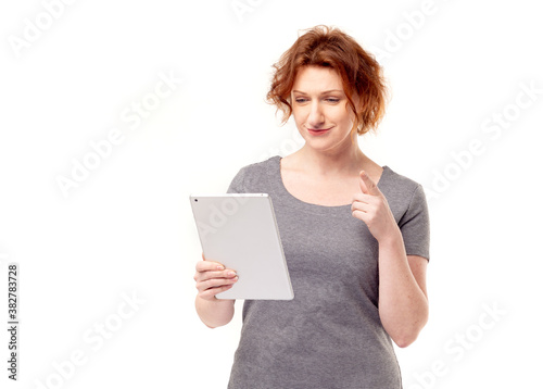Red haired adult mature woman in gray dress looking at mobile tablet in her hands 