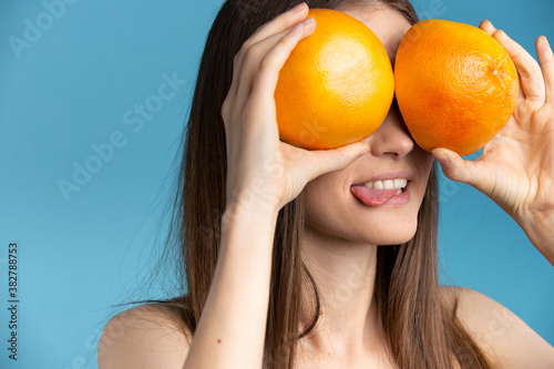 Young awesome girl holding juicy halves of grapefruit against her eyes , sticking out tongue. Blue cut out background