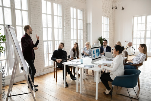 Confident businessman present business project on whiteboard for excited diverse employee at meeting or training. Male coach or trainer talk make flip chart presentation for businesspeople in office.