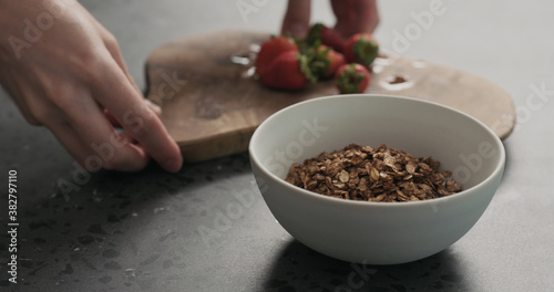 man hand place white bowl with chocolate granola, strawberries and glass for yogurt on a concrete countertop
