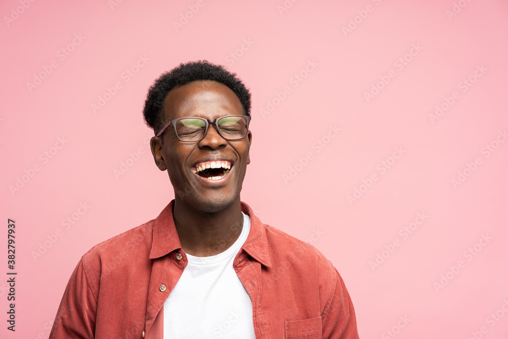 Fototapeta premium Laughing positive young Afro American man in red shirt in good mood, poses at camera with closed eyes. Overjoyed Black male in spectacles shows her broad smile, isolated on studio pink background. 