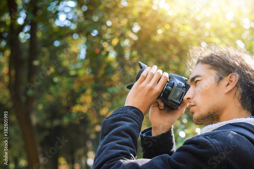 Man with dark brown curly hair in central park of the city takes pictures of colorful bright trees. Sunlight in the right upper corner, copy space on the left.