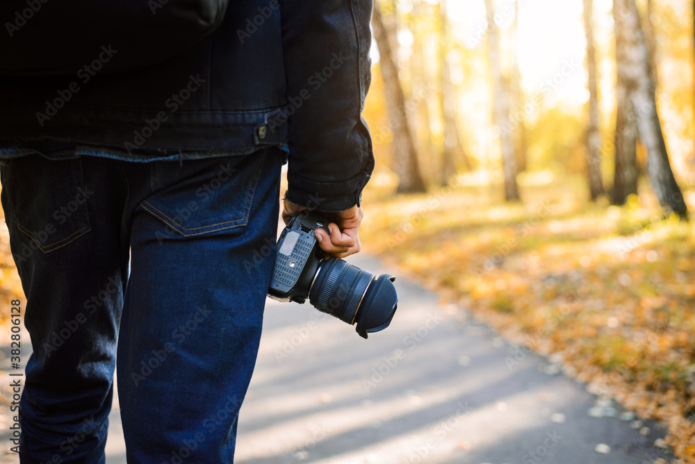 Fototapeta premium Man photographer from his back, walking in the park with camera in his hands. Autumn view on the background.