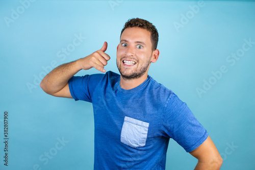 Young handsome man wearing blue casual t-shirt over isolated blue background smiling doing phone gesture with hand and fingers like talking on the telephone
