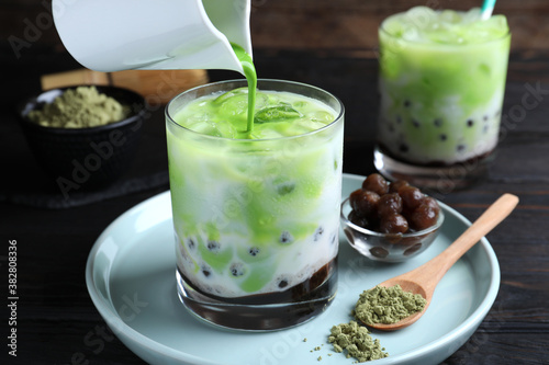 Pouring green matcha into glass with milk bubble tea at black wooden table, closeup
