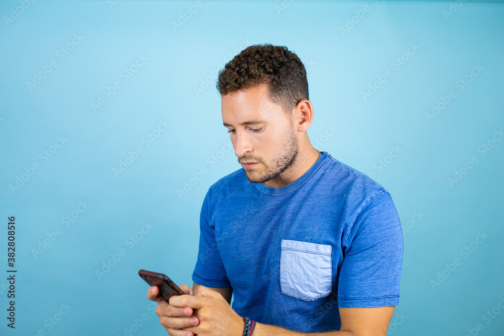 Young handsome man wearing blue casual t-shirt over isolated blue background texting with his phone Young handsome man wearing blue casual t-shirt over isolated blue background texting with his phone