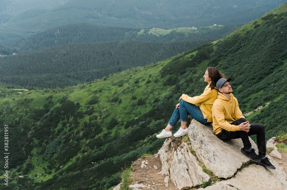 Inspirational landscape. Love in the mountains. Happy couple in love sits on the edge of the cliff back to back on the big stone with an amazing mountains view on the background