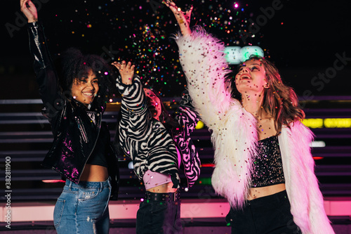 Three diverse women throwing confetti in the air at night. Happy girls celebrating outdoors in front of neon lights.