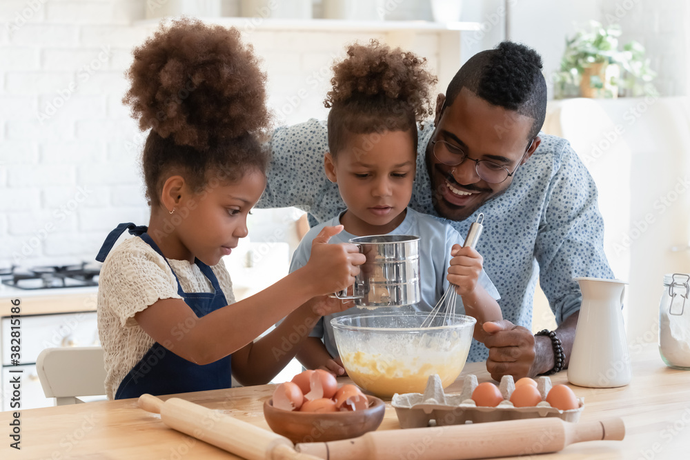 Happy young african american dad watching two adorable mixed race kids siblings preparing dough for homemade pastry, enjoying weekend family activity together in kitchen, culinary recipe concept. - obrazy, fototapety, plakaty 