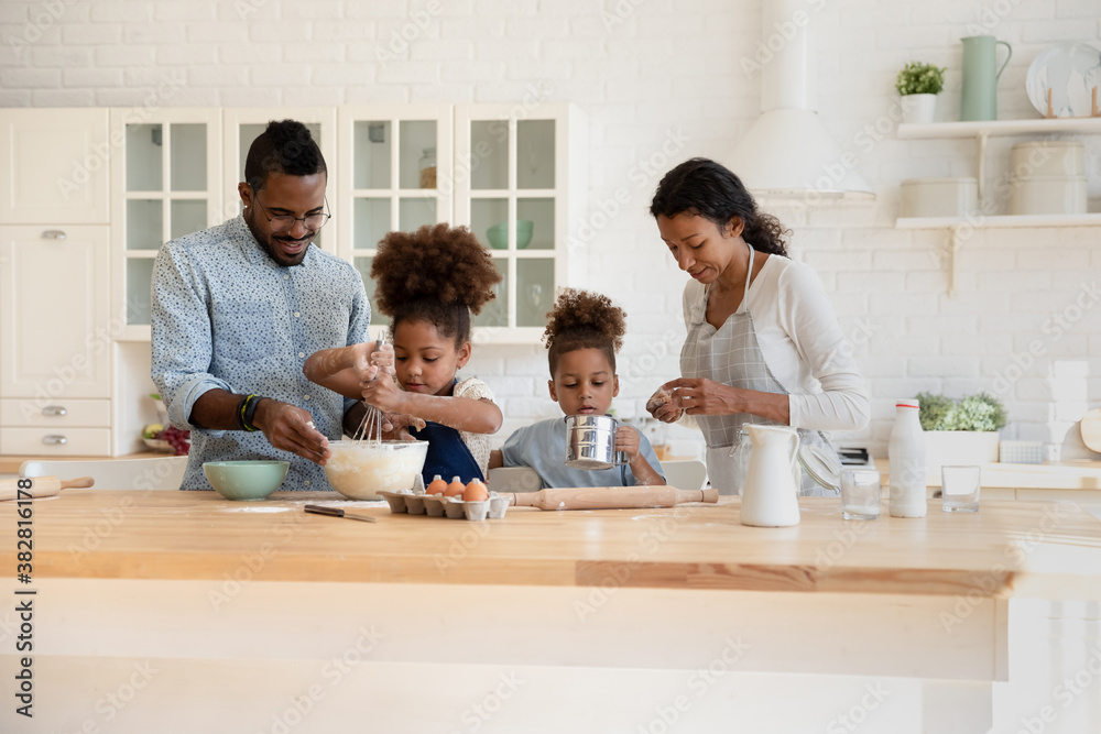 Front view happy african american full family enjoying preparing morning weekend meal, mixing ingredients for dough, cooking pancakes muffins or homemade pizza together, standing at wooden countertop. Front view happy african american full family enjoying preparing morning weekend meal, mixing ingredients for dough, cooking pancakes muffins or homemade pizza together, standing at wooden countertop.