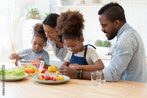 Happy mixed race young parents teaching little kids siblings cooking fresh healthy vegetarian food at home  chopping vegetables together in modern kitchen  positive bonding family weekend pastime.