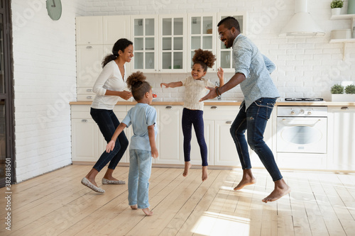 Full length overjoyed african american couple jumping with joyful energetic children siblings barefoot on warm kitchen floor, celebrating freedom weekend or enjoying stress free leisure active time.