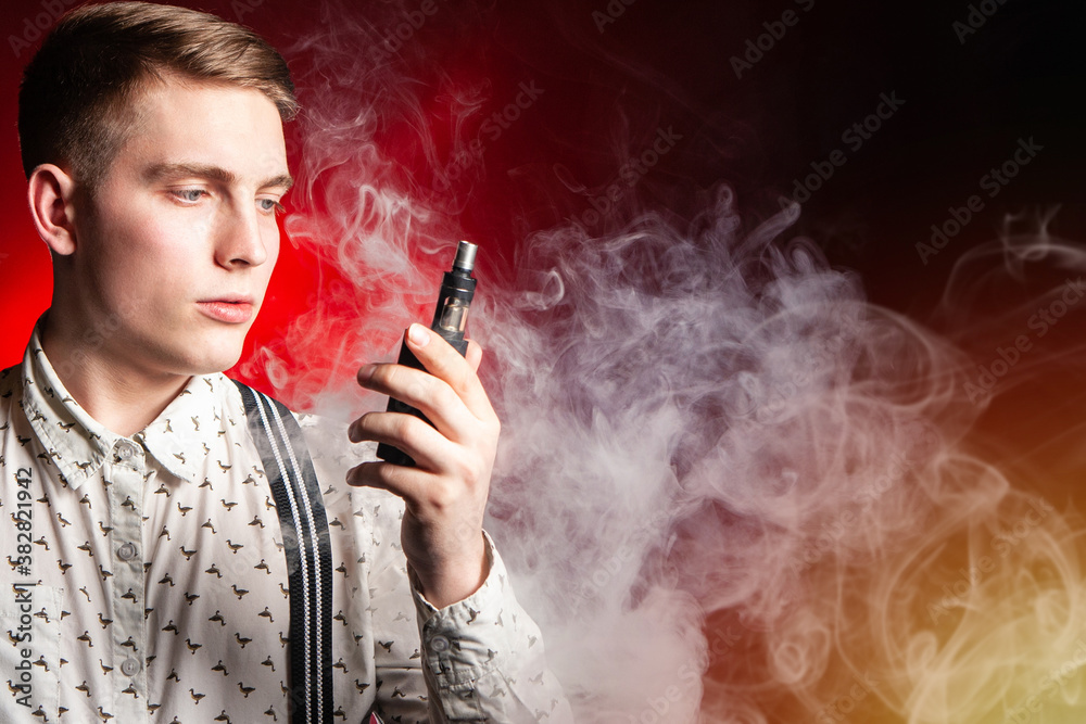 Fototapeta premium The concept of vaping in red. A young man with an electronic cigarette in his hands against the background of smoke. Space for text. VAPE shop. Smoking electronic cigarettes. The pleasure of vaping.