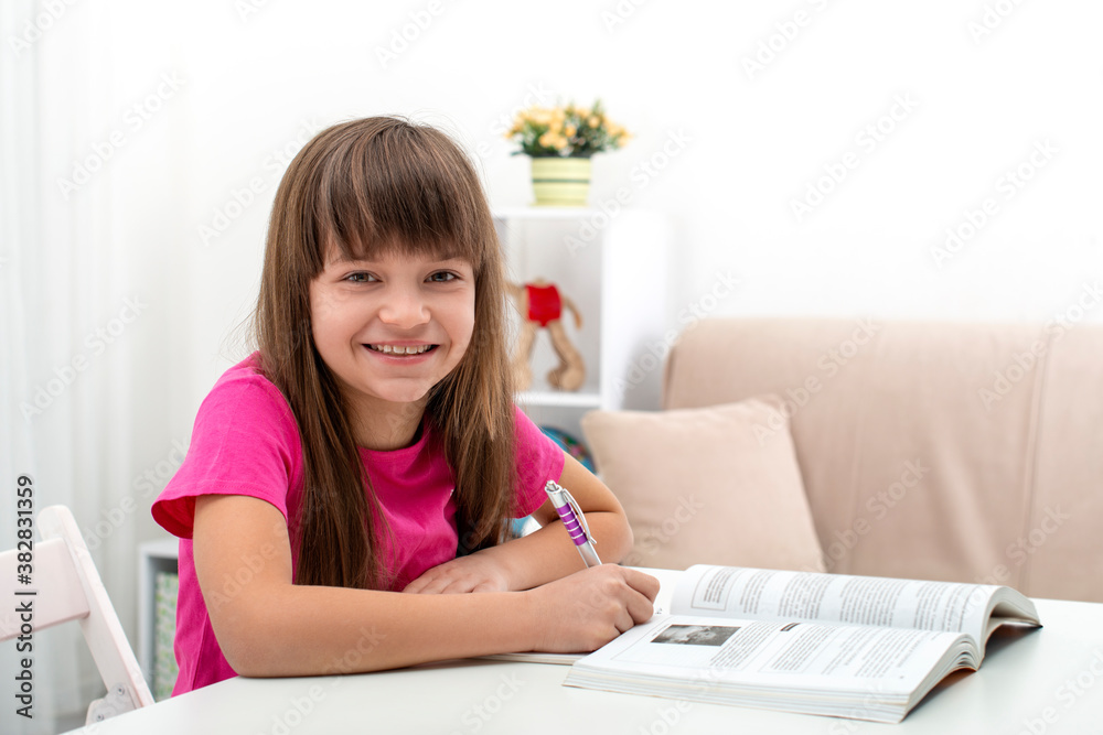 Blonde schoolgirl wearing pink shirt sits at a table at home and does homework Blonde schoolgirl wearing pink shirt sits at a table at home and does homework