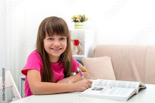 Blonde schoolgirl wearing pink shirt sits at a table at home and does homework