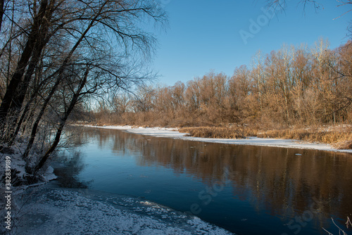 Silhouettes of trees of deciduous forest and river with stripes of snow and ice along the banks on a sunny day. . © APHOTOSTUDIO