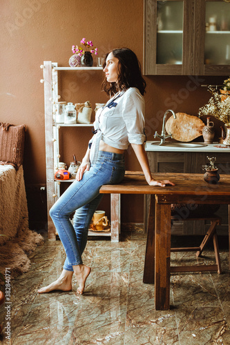 A young woman is standing in the kitchen, leaning on the table and looking out the window.