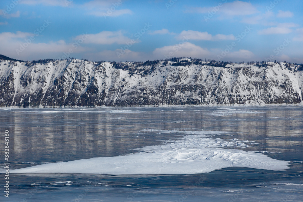 Beautiful snow formations on smooth glowing ice surface of frozen Baikal lake and covered with snow mountains on background, scenic winter landscape