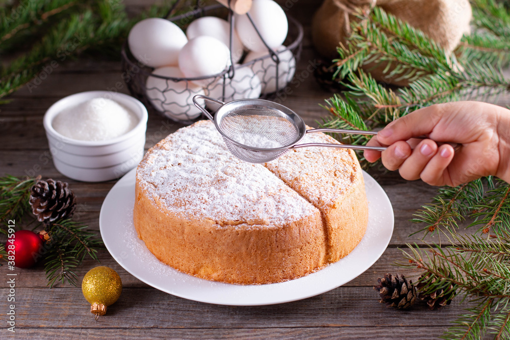Homemade sponge cake with powdered sugar on a white plate on a wooden table. New Year and Christmas sponge cake on white plate so soft and delicious with ingredients: eggs, flour, milk Homemade sponge cake with powdered sugar on a white plate on a wooden table. New Year and Christmas sponge cake on white plate so soft and delicious with ingredients: eggs, flour, milk