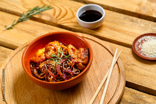 Soby with chicken and sesame seeds in teriyaki sauce in a clay plate on a wooden stand and sticks next to sesame seeds, herbs and soy sauce on a wooden table. photo