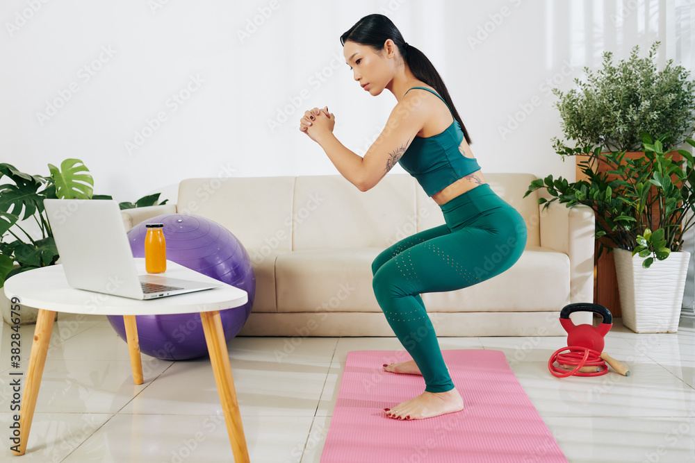 Fototapeta premium Young Asian woman working out at home, doing squats and looking at laptop screen