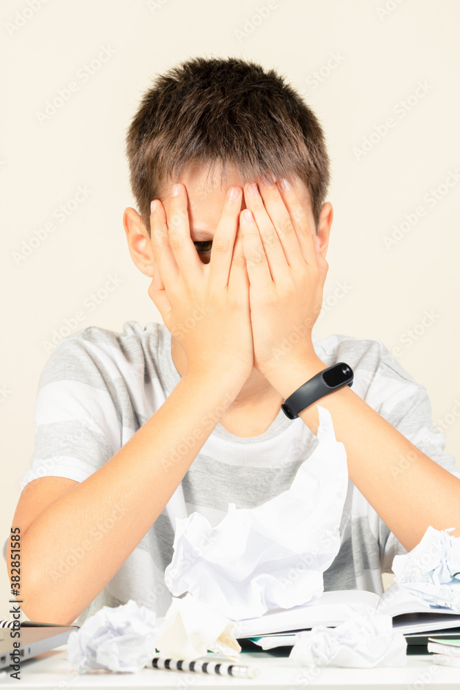 Kid covering his face with hands and peering out with one eye between fingers. Sad boy have learning problem with homework