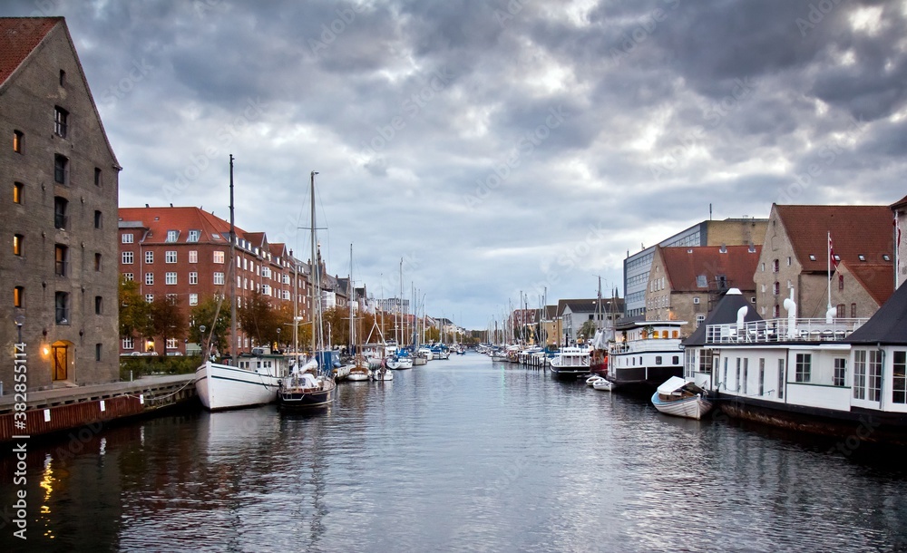 Fototapeta premium White moored motor boats in a channel and european houses on the embankment against cloudy sky in Copenhagen, Denmark.