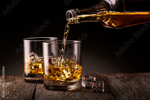 Pouring whiskey from bottle into the glass with ice cubes on black background.