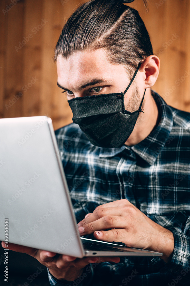 Remote operation during the quarantine, to print the text and conduct important business. A handsome young freelancer in a black protective mask works on a silver laptop at home. Remote operation during the quarantine, to print the text and conduct important business. A handsome young freelancer in a black protective mask works on a silver laptop at home.