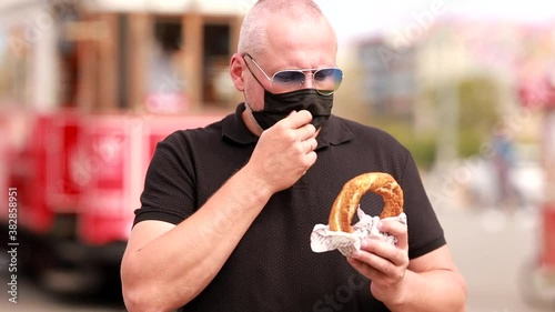 Video, an emotional adult man in a black shirt and sunglasses eats a simit bun on a city street. People are walking next to the man and a tram is passing by. On a sunny day in summer. photo