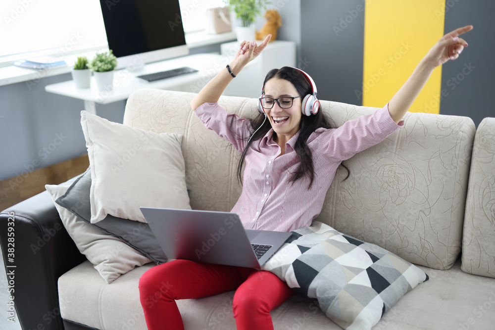 Obraz premium Beautiful woman in glasses and headphones sit on couch, listen music and dance. Gray laptop on knees and female hands are up.