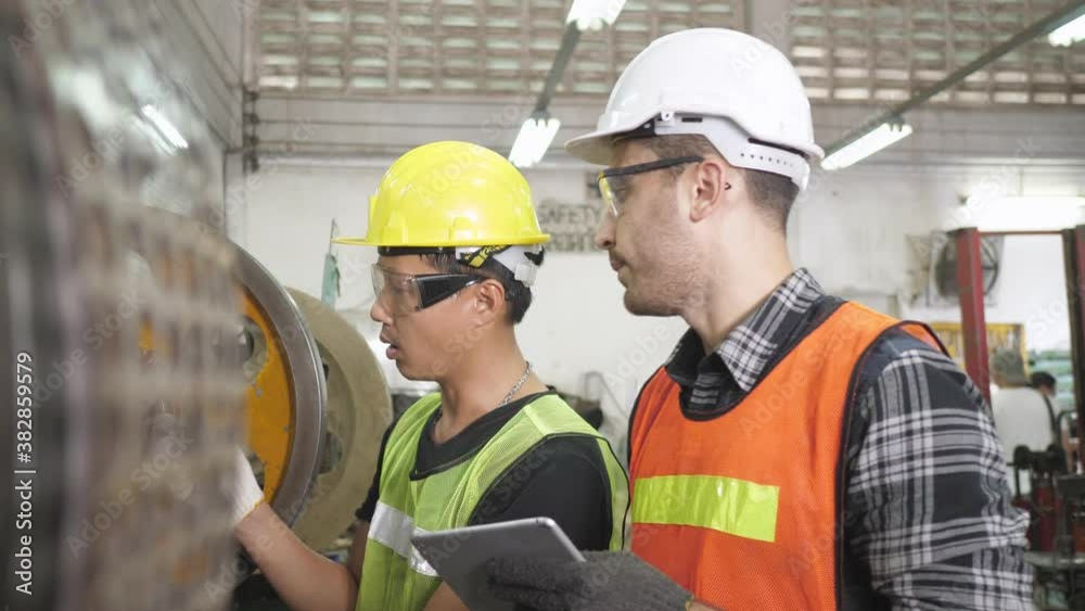 Caucasian and Asian engineer worker use tablet for maintenance checking machine. Teamwork with safety uniform in manufacturing metal sheet factory production. Dolly camera panning view from machine Caucasian and Asian engineer worker use tablet for maintenance checking machine. Teamwork with safety uniform in manufacturing metal sheet factory production. Dolly camera panning view from machine