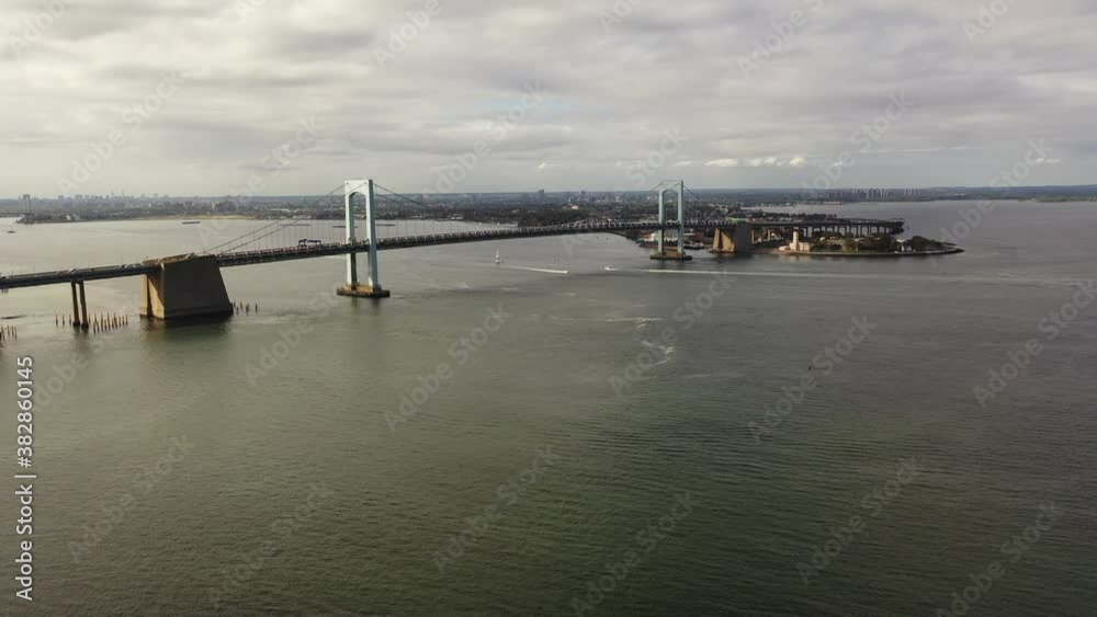 An aerial view of the Throgs Neck Bridge from over the East River. The drone dolly in towards the bridge as it pan left and boom up slowly.  It is a cloudy day & the river is calm.